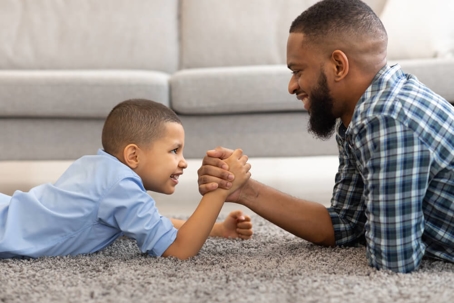 Happy Black Father And Son Armwrestling Lying On Floor Indoors